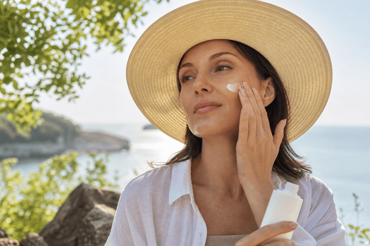 Femeie adultă aplicând cremă cu SPF pe față, într-un decor de vară, cu pălărie de soare și lumină caldă