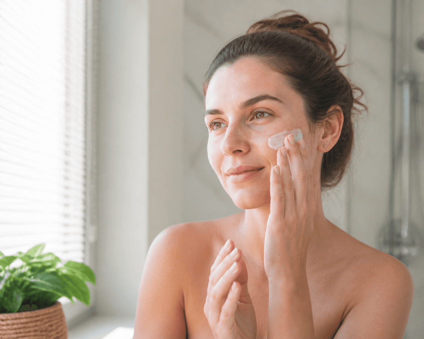 Femeie adultă aplicând o cremă cu SPF lejeră pe față, într-o baie luminoasă, într-o zi caldă de vară